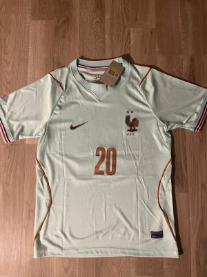 Frankrike Doue VM-2026 Fotbollströja S - Snygg ljusgrön fotbollströja med Frankrikes landslagsmärke och två stjärnor på bröstet. Bronsfärgade detaljer, nummer 20 och namnet 'DOUE' på ryggen. Tröjan har korta ärmar och randiga muddar i blått, vitt och rött. Perfekt för match eller träning. HELT NY!