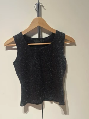 Glittrig svart crop top från B-Young - Snygg svart crop top från B-Young med glittrigt material och breda axelband. Toppen har en figurnära passform och rund halsringning. 