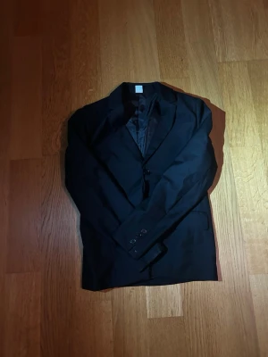 Klassisk svart kavaj - En stilren svart kavaj med två knappar framtill och klassisk krage. Kavajen har en slimmad passform och är helfodrad för extra komfort. Perfekt för dig som vill ha en enkel men snygg look till både vardag och mer uppklädda tillfällen.