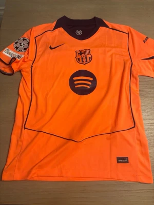 Orange FC Barcelona matchtröja - Snygg orange FC Barcelona matchtröja med mörklila detaljer, klubbmärke och Nike-logga på bröstet samt Spotify-logga. Nummer 28 och namnet Roony på ryggen. Champions League-märke på ärmen och UNHCR-tryck nedtill bak. Perfekt för fotbollsfans!