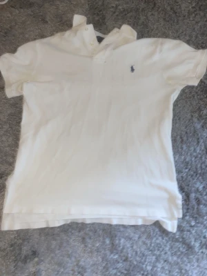 Vit pikétröja från Polo Ralph Lauren - Klassisk vit pikétröja från Polo Ralph Lauren i bomull. Tröjan har korta ärmar, knappar vid halsen och den ikoniska broderade loggan på bröstet. Perfekt för en clean och stilren look.