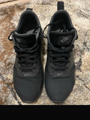Nike Air Max Tavas svarta sneakers - Säljer ett par svarta Nike Air Max Tavas sneakers med klassisk swoosh-logga. Skorna har mesh- och syntetmaterial, svarta skosnören och en låg siluett. Perfekta för dig som gillar stilrena och sportiga sneakers.