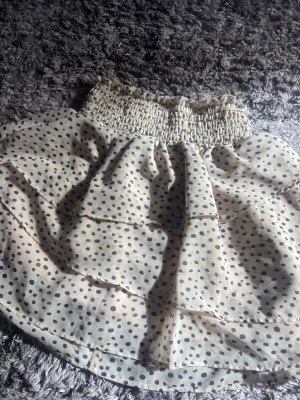 Beige prickig volangkjol - Supersöt beige/vit kortkjol med svarta prickar och bred smock i midjan. Kjolen har flera volanger som ger en luftig och lekfull känsla. Liten storlek men den är naturligt lång. 