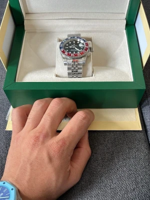 Silverfärgad klocka med röd och blå bezel - Stilren klocka i silverfärgat metallarmband och boett. Urtavlan är svart med tydliga markeringar och visare, samt en bezel i rött och blått som ger en sportig känsla. Klockan levereras i en grön och vit box.