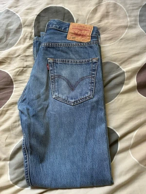 Levi's 501 ljusblå raka jeans - Klassiska Levi's 501 jeans i ljusblå denim med rak passform och fem fickor. Jeansen har den ikoniska läderlappen bak i midjan och röd Levi's-tagg på bakfickan. Perfekta för en avslappnad och tidlös stil. W34 L32