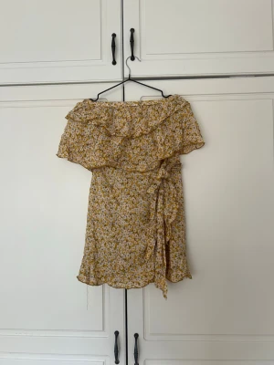 Gul blommig omlottklänning/ blustopp från Commodo - Söt gul kort omlottklänning från Commodo med blommigt mönster, volangdetaljer och offshoulder-design. Perfekt för varma dagar och har ett knyt i midjan för extra passform. Lätt och luftig känsla.