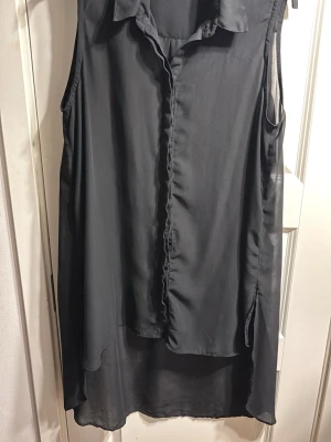 Svart ärmlös skjortklänning - Svart ärmlös skjortklänning i ett tunt, lätt material med krage och knappar framtill. Klänningen har en lös passform och är något längre baktill. Perfekt för en stilren och enkel look.