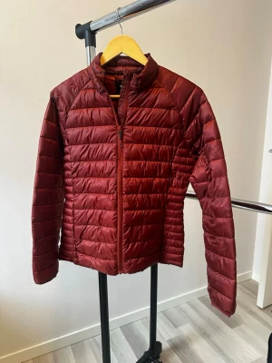 Roströd pufferjacka med quiltad design - Snygg vinröd pufferjacka med horisontella quiltade sömmar från lager 157. Jackan har dragkedja framtill och är tillverkad i ett glansigt, lätt material som håller värmen. Perfekt för kalla vinterdagar och ger en stilren look. Helt ny aldrig använd.