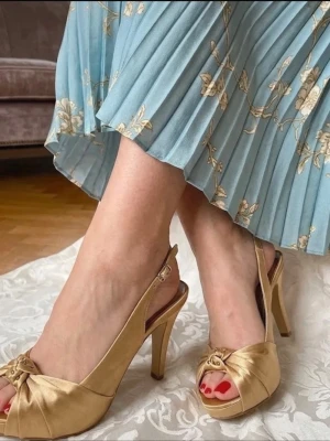 Guldiga klackskor från Luana - Eleganta guldiga klackskor från Luana med öppen tå och dekorativ rosett framtill. Skorna har en smal rem runt hälen och en stilren klack som ger en sofistikerad look. Perfekta för speciella tillfällen. Som student eller annat 