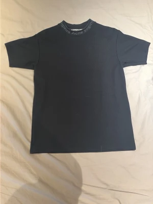 Svart t-shirt från Acne Studios - Svart t-shirt från Acne Studios med ribbad hög krage där Acne Studios-loggan är invävd runt om. Klassisk passform och korta ärmar. Perfekt för dig som gillar stilrena och trendiga plagg.
