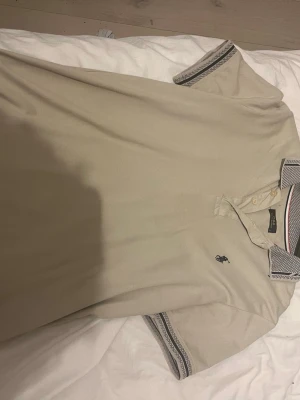 Polo Piké  - Snygg beige pikétröja med korta ärmar och broderad logga på bröstet. Kragen och ärmsluten har randiga detaljer i svart och vitt. Perfekt till jeans eller shorts för en clean look.