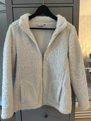 Vit teddyjacka från Isolde - Mysig vit teddyjacka från Isolde i storlek M. Jackan har dragkedja framtill, två fickor och hög krage. Tillverkad i fluffigt teddy-material som ger en trendig och avslappnad look.