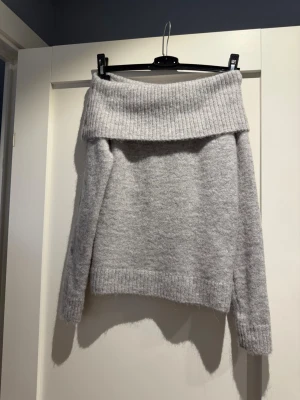 Ljusgrå off shoulder stickad tröja - Mysig ljusgrå stickad tröja från Gina Tricot med bred ribbad off shoulder-krage. Tröjan har långa ärmar och ribbade muddar vid ärmslut och nederkant. Perfekt för kalla dagar och enkel att styla till olika outfits.