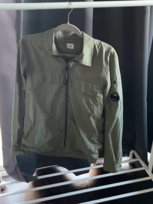 CP Company jacka - Snygg CP Company jacka i en stilren olivgrön färg. Jackan har dragkedja framtill, flera praktiska fickor och den klassiska CP Company-linsen på ärmen. Perfekt som lättare jacka för vår och höst. 