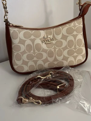 Coach beige och brun handväska - Snygg handväska från Coach i beige canvas med klassiskt monogrammönster och bruna läderdetaljer. Väskan har guldfärgade metalldetaljer och medföljer en avtagbar axelrem i brunt läder. Perfekt accessoar för en trendig look. PRIS KAN DISKUTERAS!