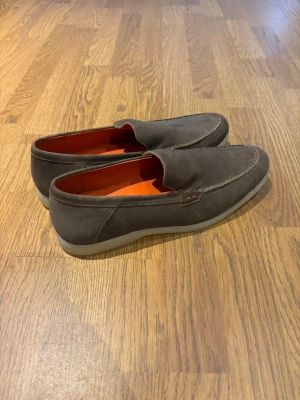 Bruna loafers med orange innersula - Snygga bruna loafers i mocka med kontrasterande orange innersula och ljus sula. Skorna har en klassisk slip-on design och diskreta sömmar längs ovandelen. Perfekta för dig som vill ha en stilren och bekväm sko med en cool detalj.