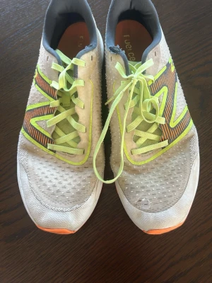 Vita sneakers med neongula detaljer - Säljer ett par vita sneakers med neongula och orange detaljer samt grå insida. Skorna har meshmaterial för bra ventilation och en sula i orange gummi med mönster för bra grepp. Snygg design med stora färgade ränder på sidan och matchande neongula skosnören.
