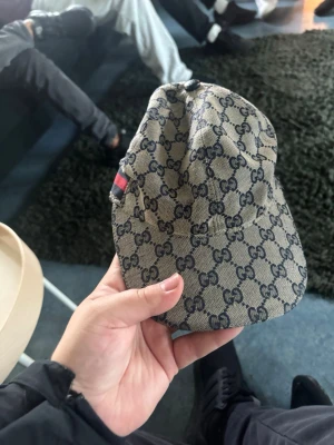 Beige Gucci keps med GG-mönster - Säljer en beige keps från Gucci med det klassiska GG-monogrammet i mörkblått över hela kepsen. Kepsen har en böjd skärm och en liten röd-grön detalj på sidan. Perfekt för dig som vill ha en stilren och lyxig accessoar.