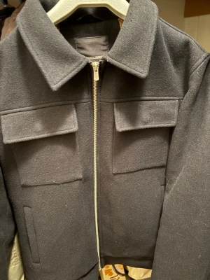 Svart overshirt med fickor och dragkedja - Stilren svart overshirt med två stora bröstfickor och dragkedja framtill. Overshirten har klassisk krage och är gjord i ett tjockare material som ger en snygg och strukturerad look. Perfekt att bära över en t-shirt eller hoodie.