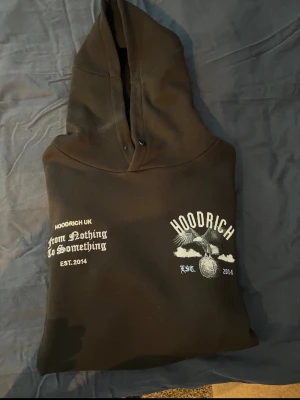 Svart hoodie från Hoodrich - Svart hoodie med tryck framtill från Hoodrich. På bröstet står det 'From Nothing To Something' och 'Hoodrich' med en örn och bollmotiv. Hoodien har huva och dragsko. Perfekt för en streetwear-look.