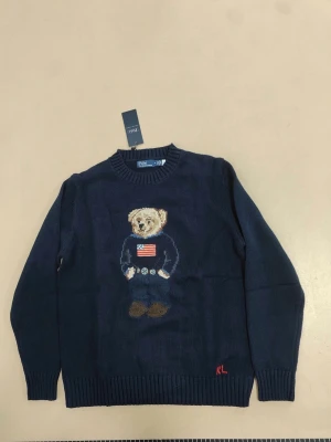 Ralph lauren polo bear blå - Mörkblå stickad tröja från Polo Ralph Lauren med ikonisk björn på bröstet, klädd i amerikansk flaggtröja och jeans. Rund halsringning och ribbstickade muddar. RL-broderi i rött nedtill. Perfekt för dig som gillar klassisk stil