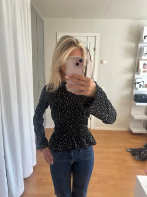 Svart prickig blus med volang Gina tricot  - Trendig svart blus med vita små prickar och smockad midja som ger en snygg siluett. Blusen har långa ärmar och volang nedtill för en feminin touch. Jättebra skick! 