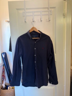 Mörkblå skjorta från Ralph Lauren - Klassisk mörkblå skjorta från Ralph Lauren med knappar framtill och liten broderad logga på bröstet. Skjortan har lång ärm och normal passform, perfekt till jeans eller chinos. Tillverkad i mjuk bomull för en bekväm känsla.
