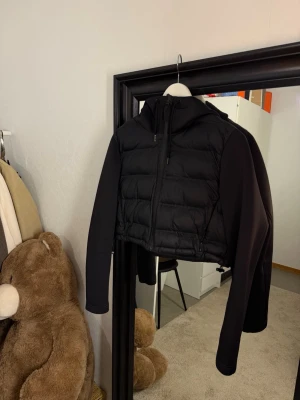 Jacka Zara  - This is a black, cropped puffer jacket with a hood and long sleeves. The body of the jacket is quilted and padded, while the sleeves and hood appear to be made from a different, smoother fabric. The jacket has a front zipper and a sporty, modern look. Funkar för S/M då man kan dra åt den 