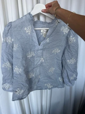 Blåvit blus med broderade blommor - Söt blåvit blus från Cream med tunna ränder och vita broderade blommor. Blusen har puffiga långärmar, lätt rynkad halsringning och en liten v-ringning framtill. Perfekt till jeans eller kjol för en fräsch look.