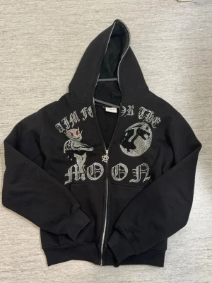 Svart hoodie med strassdetaljer - Snygg svart hoodie med dragkedja och huva. Framsidan har coola strassdetaljer med texten 'Aim for the moon' och motiv av en drake och måne. Hoodien har ribbade muddar vid ärmar och nederkant. Perfekt för dig som gillar streetwear och statement-plagg.