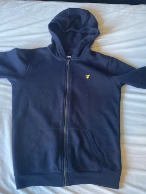 Mörkblå lyle & Scott zip-up hoodie  - Mörkblå hoodie med dragkedja och huva. Har två fickor framtill och en liten gul logga på bröstet. 