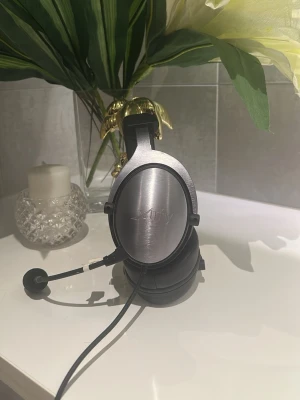 Xtrfy H1 Gaming Headset kostar 1175kr - Snygga svarta over-ear hörlurar med mikrofon och kabel. Hörlurarna har en matt finish och rundad form med metallfärgade detaljer på kåporna. Perfekta för gaming, streaming eller samtal. Bekväm vadderad bygel och justerbar mikrofonarm.