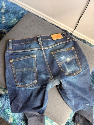 Nudie jeans Grim tim - Säljer ett par mörkblå jeans med tydliga slitningar och kontrastsömmar. Jeansen har raka ben, två bakfickor med dekorativa sömmar och en klassisk läderpatch i midjan. Materialet är kraftig denim som ger en cool vintagekänsla.  W33 L32, midja 43cm, längd 106.5 cm, benöppning 17.5cm