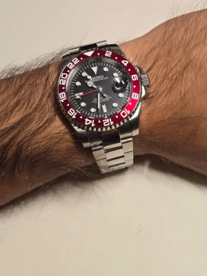 Seiko GMT red mod - Helt ny Seiko GMT Red mod som är väldigt stilren och sticker ut med sin helröda GMT-bezel 🔴✨  Boetten är 39 mm och ger en riktigt bra passform på handleden ⌚️ Svart urtavla tillsammans med den röda bezeln ger klockan en riktigt snygg look 🔥 Rostfritt stål och datumvisning med förstoringsglas ✅ GMT-visare för extra tidszon 🌍  En riktigt snygg och unik klocka för dig som gillar Seiko mods ✨  Skickas välpackad 📦 Kontakta gärna för fler bilder eller mer info!