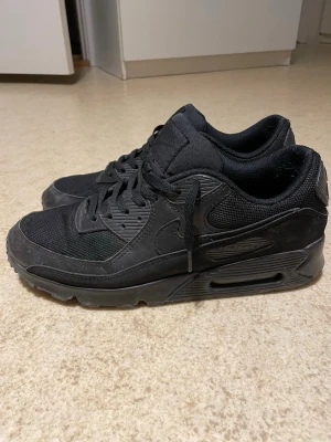 Airmax 90 svarta - Säljer ett par helsvarta Nike Air Max 90 sneakers med klassisk siluett. Skorna har ovandel i mesh och mocka, samt detaljer i plast och gummi. Tjock sula med synlig Air-enhet och Nike-logga på sidan. Perfekta för dig som gillar streetwear och sportig stil.