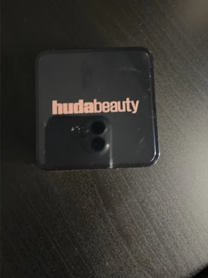 Huda Beauty Easy Bake  duo - Säljer en Huda Beauty Easy Bake ögonskugga duo i en kompakt fyrkantig förpackning. Innehåller två nyanser: en ljus beige och en ljus rosa, perfekta för att skapa mjuka och naturliga ögonlooks. Praktisk och stilren design med genomskinlig botten.