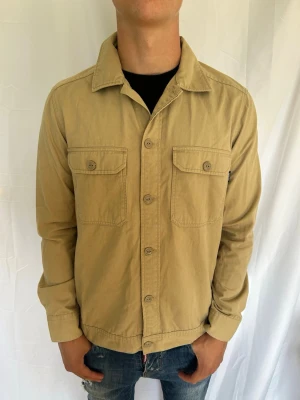 Overshirt - Stilren beige overshirt med knäppning framtill och två bröstfickor med lock. Skjortkrage och raka ärmar. Tillverkad i ett mjukt bomullstyg som ger en avslappnad look. Perfekt att bära öppen över en t-shirt.
