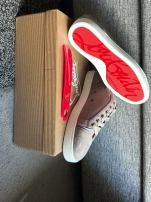 Gråa sneakers med röd sula - Snygga sneakers i grå mocka med klassisk röd sula och vita kanter. Skorna har rund tå, matchande grå skosnören och diskreta sömmar. Perfekta för dig som vill ha en stilren men ändå unik look.