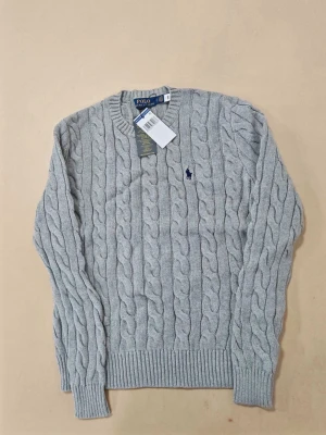 Grå kabelstickad tröja Polo Ralph Lauren - Snygg grå kabelstickad tröja från Polo Ralph Lauren med rund halsringning och ribbade muddar. Tröjan har det klassiska lilla mörkblå logobroderiet på bröstet och är tillverkad i mjukt material. Perfekt för dig som gillar stilrena och tidlösa plagg.