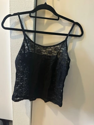 Svart spetslinnetopp med smala band - Snygg svart linnetopp i spets med smala axelband och transparenta detaljer. Toppen har ett feminint mönster och är perfekt för att styla lager på lager eller till ett par jeans. Croppad passform och mjukt material.
