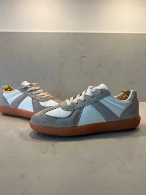 Vita och grå sneakers med gummisula - Snygga sneakers i vitt och ljusgrått med ovandel i mocka och textil. Skorna har klassisk låg profil, vita snören och en brun gummisula som ger retrovibbar. Perfekta för dig som gillar stilrena och tidlösa sneakers.