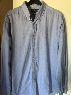 Blå skjorta från Polo Ralph Lauren - Klassisk ljusblå skjorta från Polo Ralph Lauren med button-down krage och broderad logga på bröstet. Skjortan har långa ärmar, knappar framtill och är tillverkad i bomull. Perfekt till en avslappnad men ändå stilren look.