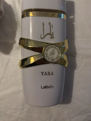 Yara parfym från Lattafa - Snygg parfymflaska i vitt med gulddetaljer och elegant design från Lattafa. Flaskan har arabiska bokstäver och texten 'YARA' på framsidan. Dekorativa guldband ger en lyxig känsla och flaskan känns modern och unik.