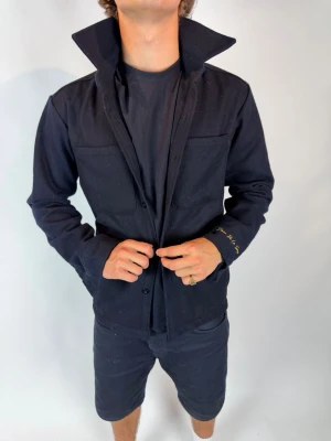 Svart overshirt med hög krage - Stilren svart overshirt med hög krage och knäppning framtill. Två bröstfickor ger en clean look och materialet ser ut att vara mjukt och lite tjockare, perfekt för lager på lager. Diskret broderad text på ena ärmen för en unik touch.