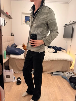 Grå camouflage vindjacka - Snygg grå vindjacka med camouflage-mönster och knappar framtill. Jackan har flera fickor, klassisk krage och långa ärmar. Perfekt för dig som vill ha en streetwear-inspirerad look. Färgen är lite oklar, den är grå men har en hint av lite mintgrönt i sig. 