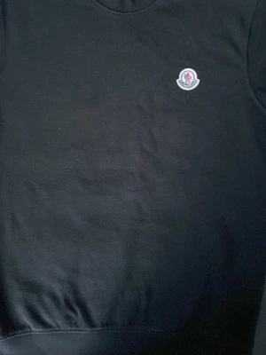 Svart sweatshirt från Moncler - Svart sweatshirt från Moncler med rund halsringning och klassisk logotyp på bröstet. Tröjan har en enkel och stilren design med ribbade muddar vid ärmslut och nederkant. Perfekt för dig som gillar exklusiva streetwear-plagg.