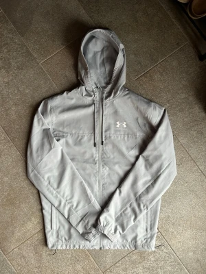 Under Armour Windbreaker - Snygg grå vindjacka från Under Armour med huva och dragkedja framtill. Jackan har elastiska muddar vid ärmsluten, två sidofickor och justerbar nederkant. Tillverkad i lätt och tunt material, perfekt för blåsiga dagar. Diskret logga på bröstet.