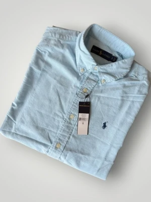 Ljusblå skjorta från Ralph Lauren - Klassisk ljusblå skjorta från Ralph Lauren med button-down krage och broderad logga på bröstet. Skjortan har långa ärmar, vita knappar och är tillverkad i mjuk bomull. Perfekt för en stilren och avslappnad look.