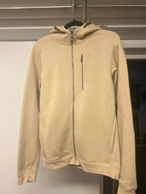 Beige hoodie från Sail Racing - Snygg beige hoodie från Sail Racing med dragkedja framtill och en extra bröstficka med zip. Tillverkad i mjukt material och har huva med dragsko. Perfekt för chill dagar eller när du vill ha en clean look. På bild 3 ser du ända defekten som knappast är synlig använd max 2 ggr