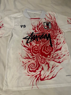 Vit Japan-tröja med röd drakdesign - Säljer en vit sporttröja med unik röd drakmönsterdesign, Japan-emblem på bröstet och svart Stüssy-tryck framtill. Tröjan har korta ärmar, klassisk krage och är tillverkad i ett lätt, andningsbart material perfekt för träning eller match.